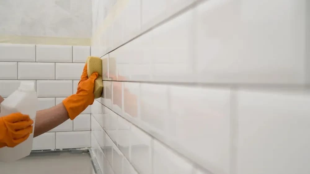 Gloved hand cleaning a tiled wall with a sponge